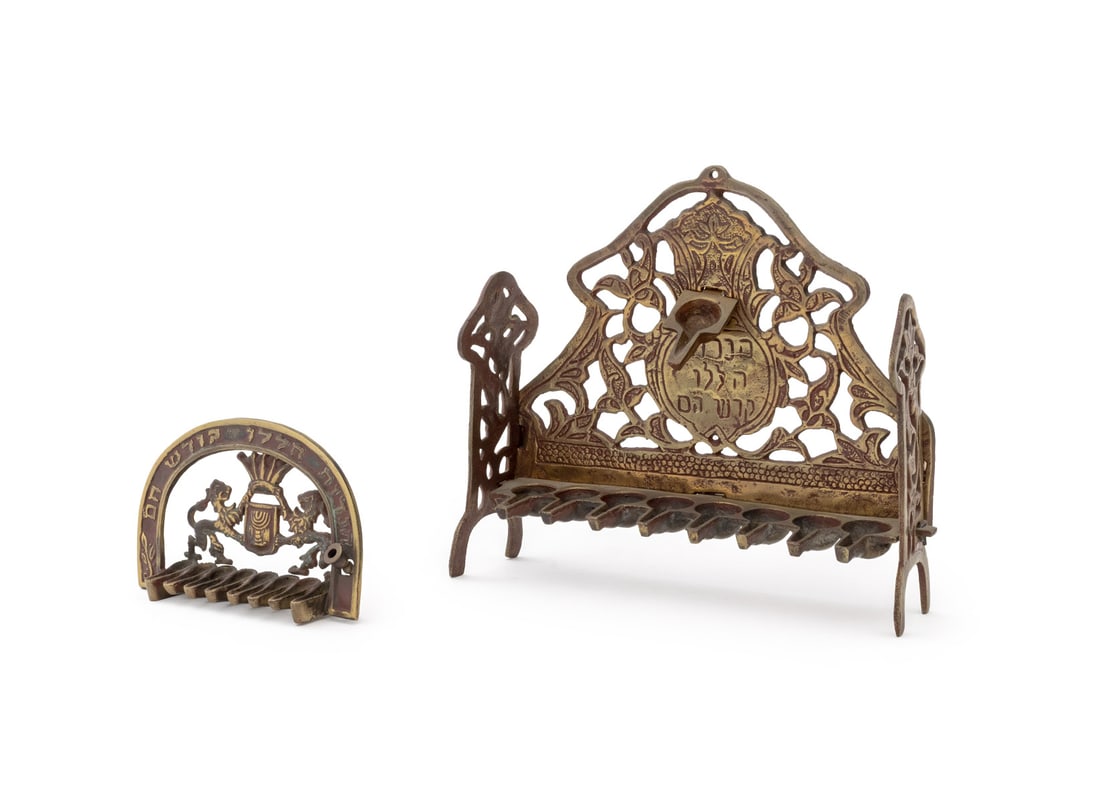 Judaica Brass Oil Lamp Menorahs, 2: Two Judaica Brass Oil Lamp Menorahs, the smaller with Lion of Judah motif below an arch with prayer in Hebrew, the larger with reticulated scrolling foliate motif. Larger: 8.75" H x 9.75" W x 2.75" D.