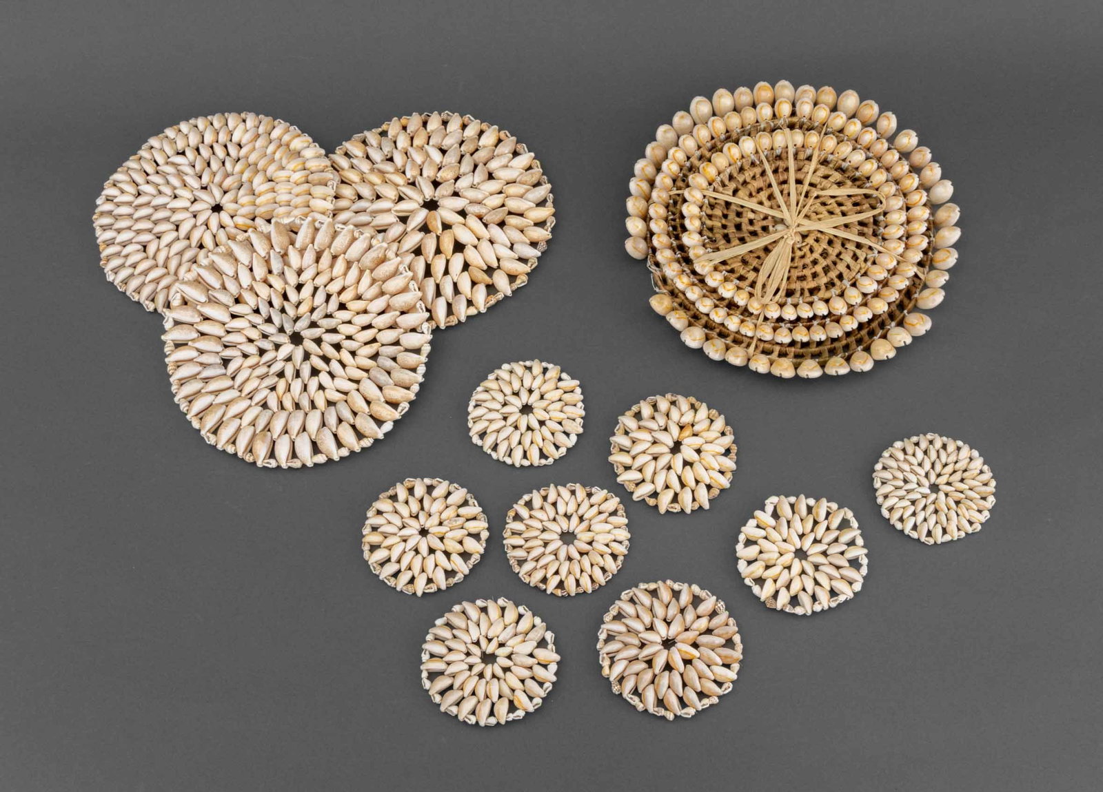 Cowrie Shell Placemats and Coasters, 14: Collection of Cowrie Shell Place Mats and Coasters, comprising: set of eight coasters, three shell placemats, and three graduated woven placemats with shell borders. Largest: 11.5" Diameter x 1" D.