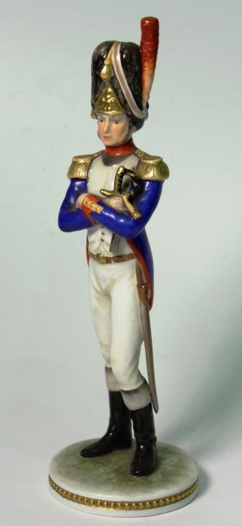 German Porcelain Figure of a Napoleonic Soldier: The elegant warrior wearing a tall bearskin shako with cords and a red side plume, gold fringed epaulettes on his navy blue cutaway, white breeches, and riding boots, in his hand a sword, the undersid