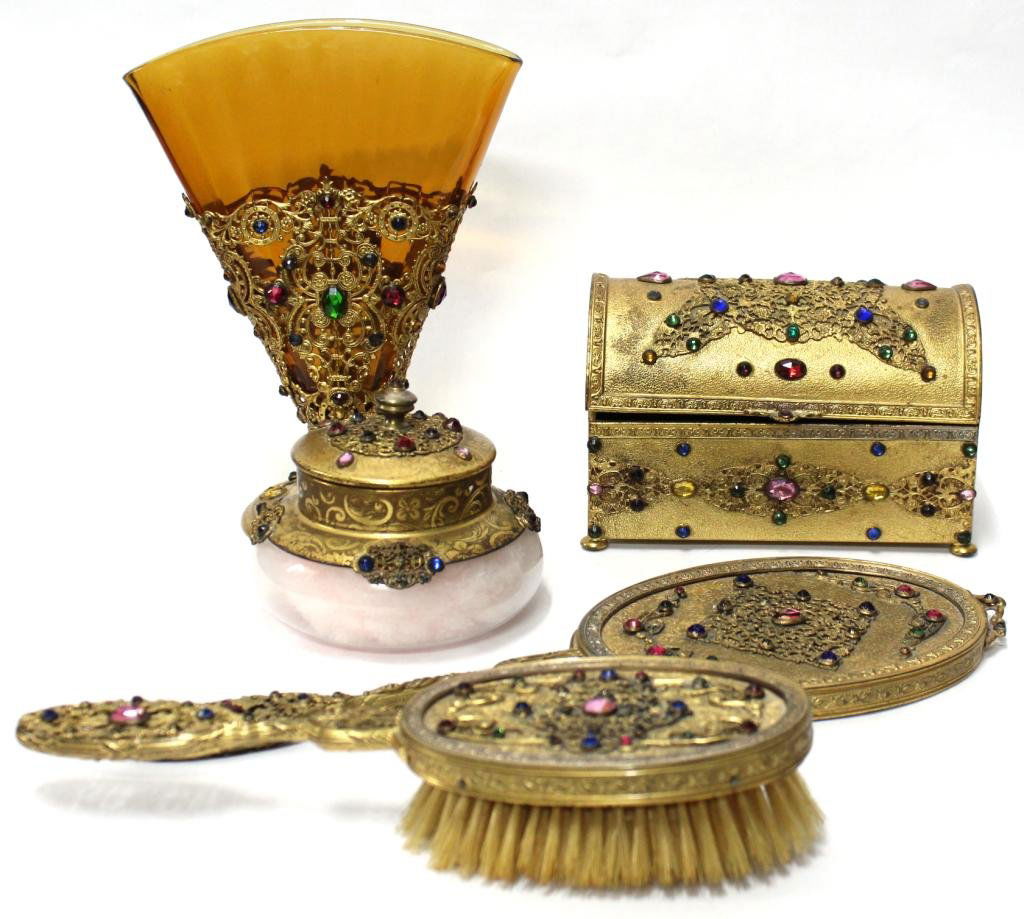 Apollo Studios Filigree & Faux Jewel Vanity Set: 1940s, comprising a brush, mirror, loose powder jar with pink swansdown puff, a jewel casket, and a fan-shaped vase with amber glass insert, the ornate gold-tone filigree metal studded with faux gems