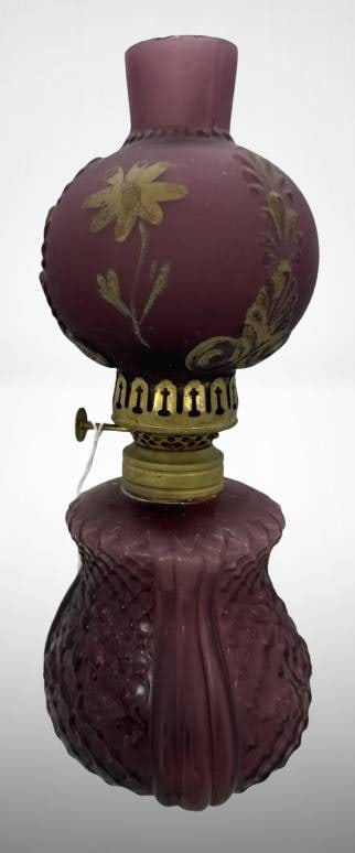 Miniature Victorian Oil Lamp: Miniature amethyst glass Victorian oil lamp featuring embossed design. ? Shade features floral patterns in gold paint. The top rim of the glass shade shows chigger bite roughness. The lamp includes a
