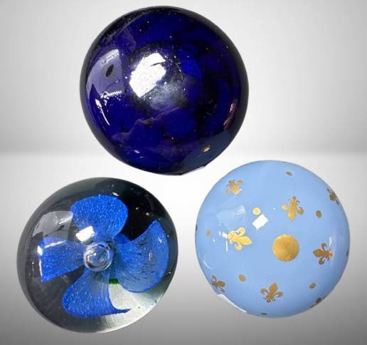 Three Glass Paperweights, 1-mrkd. Baccarat: This lot includes (3) glass paperweights with varying designs. One is marked Baccarat on light blue glass adorned with golden fleurs-de-lis. The other pieces feature a cobalt-blue sphere and a clear g