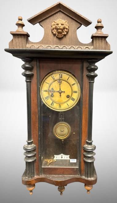 Ornate Wooden Wall Clock with Lion's Head: This ornate wooden wall clock showcases a carved lion's head at the pediment, a brass pendulum, and Roman numeral dials beneath a glass panel. Mechanically driven, it includes a key and has visible in