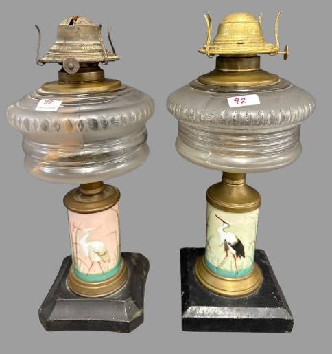 Glass Oil Lamps with Bird Motif Bases: This pair of oil lamps features hand-painted bases adorned with bird motifs, one depicting a white heron and the other a black stork. Each has ribbed glass globes and metal burners. Measure 11.5". No
