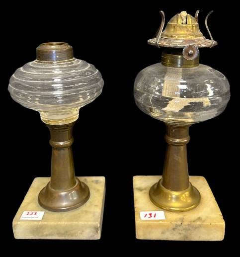 (2) Oil Lamps: This lot features (2) clear glass oil lamps, each mounted on a brass stem and marble base. One retains its complete burner assembly, highlighting functional and historical detail. Both measure approx.