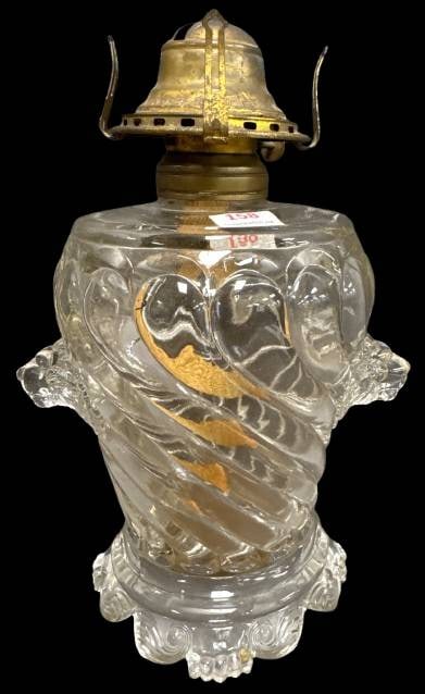 Clear Glass Victorian-era Oil Lamp with Lion Head Details: A clear glass oil lamp featuring a swirl pattern base with lion's head handles. The brass burner appears intact, and the piece exhibits no chips, cracks, or repairs. It is a preserved item with unique
