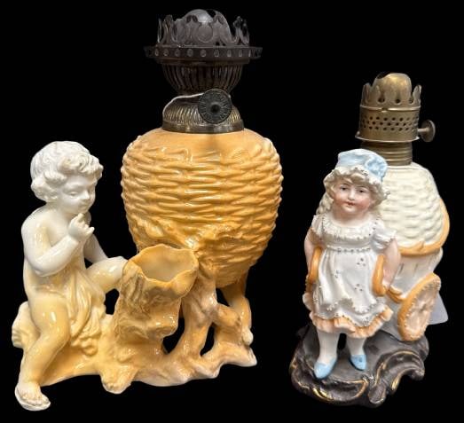 Ceramic Figural Oil Lamps: This lot includes (2) figural porcelain oil lamps: one with a child seated by a tree and another with a girl pulling a cart. Each lamp features a metal burner. Please note some rough spots present but