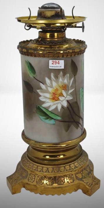Brass and Porcelain Oil Lamp With Floral Design: This brass oil lamp features a detailed floral design on painted glass, depicting white lilies and green leaves. The base includes stamped decoration, while the burner is marked with “AGUILA.”
