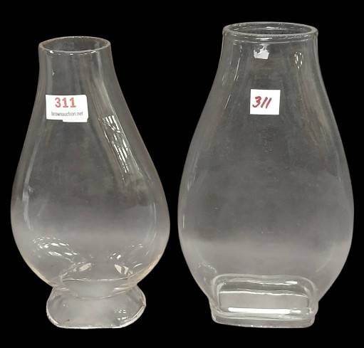 (2) Glass Lamp Chimneys: This lot includes (2) glass lamp chimneys in different shapes and bases and meas. 6.5" and 7" tall. Each piece bears signs of use and wear, with visible surface imperfections. In-house shipping availa