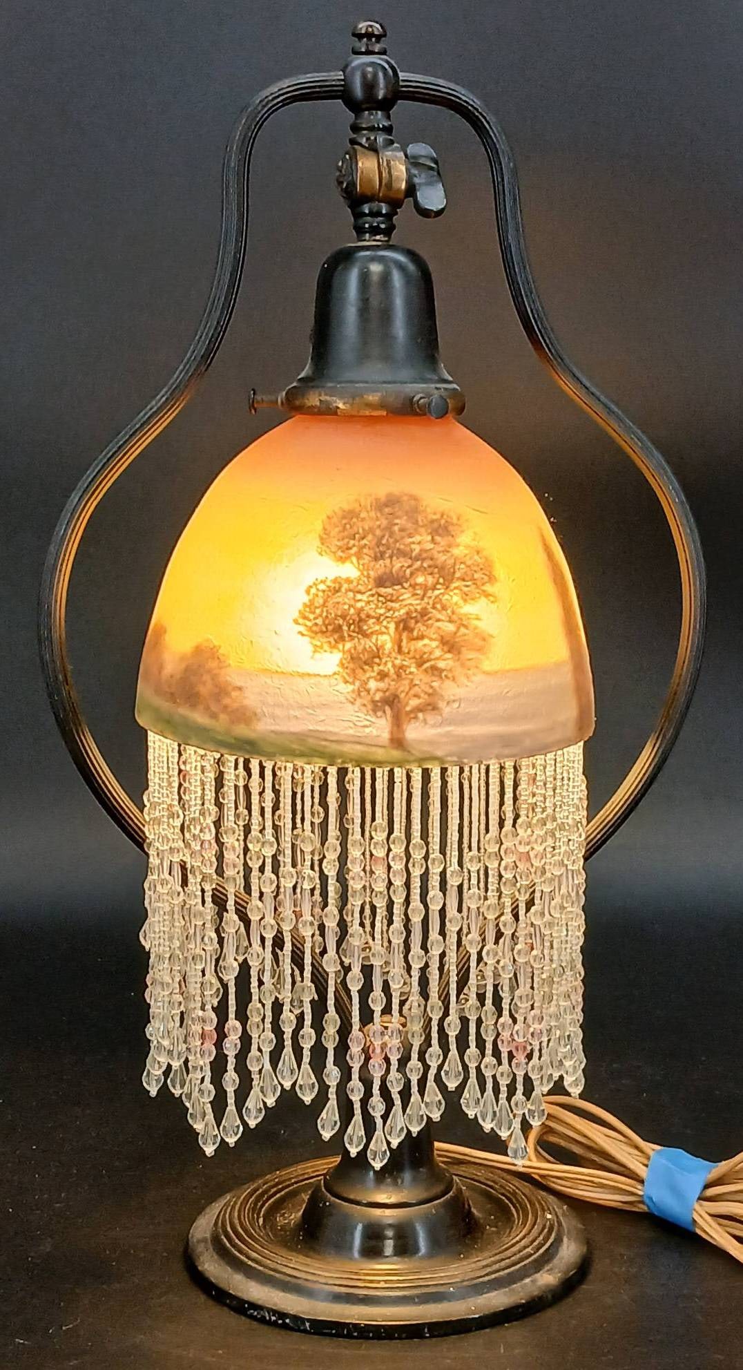 Hand-Painted Glass Beaded Boudoir Lamp: Hand-painted glass boudoir lamp with a sunset landscape theme, featuring cascading beaded trim and metal accents. Light wear and scratches are visible, including a large crack on the shade. Mark on th