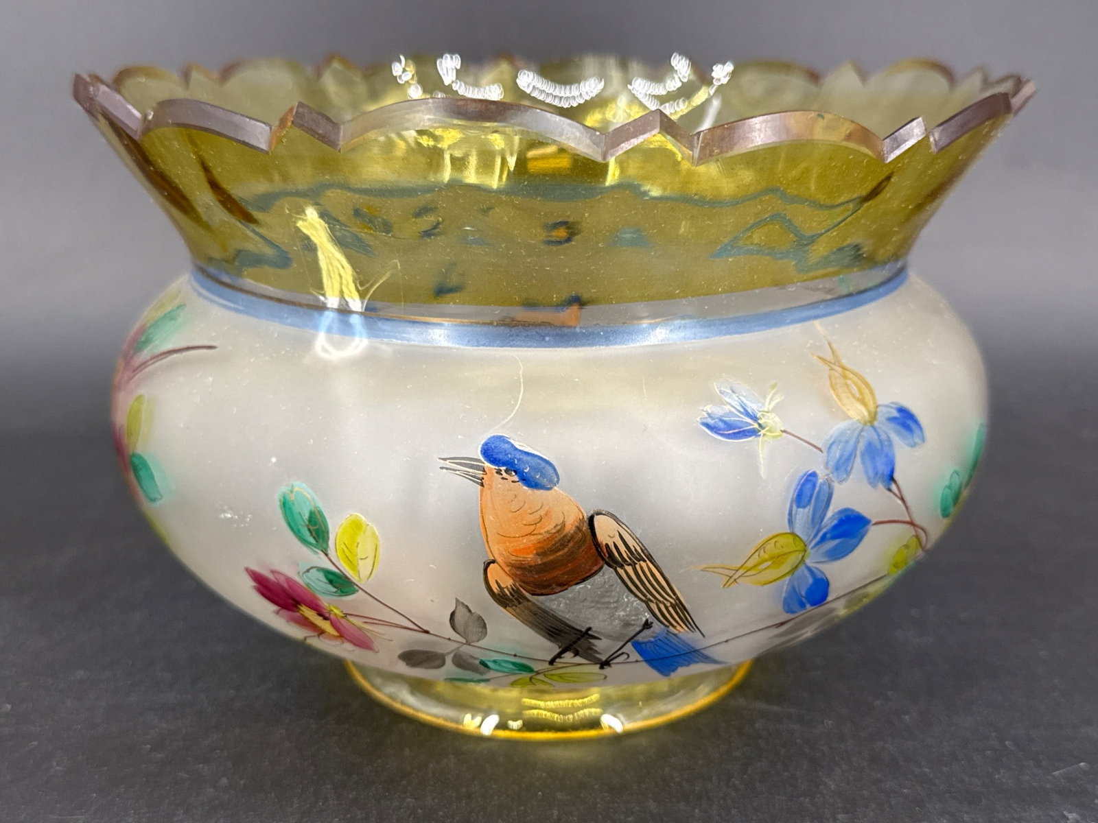 Hand-Painted Glass Bowl With Bird Motif: A hand-painted glass bowl featuring colorful bird and floral motifs. Measures approximately 4" in base diameter. Notable chip on the base. Elegant scalloped edge with gold detailing. In-house shipping