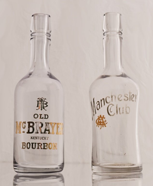 Whiskey Advertising Enamel Label Back Bar Bottles: Two Whiskey Back Bar Bottles. One for Manchester Club which is Enamel label and the other for Old McBrayer Kentucky Bourbon which is etched gold. It has a crescent scratch on the top. The Manchester h