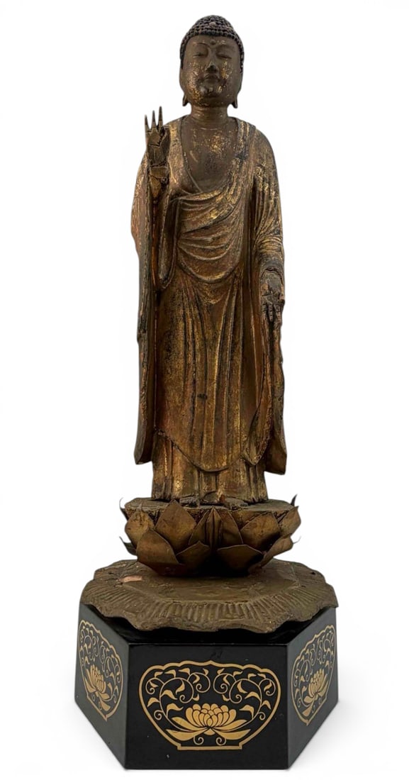 East Asian Gilt Wood Standing Buddha Figure: Carved wood standing Buddha with gilt surface, blue-painted hair, raised hand gesture, and lotus-form base, mounted on later black lacquered stand. Approximately 16.75H x 5.93W x 4.72D in. Measurement