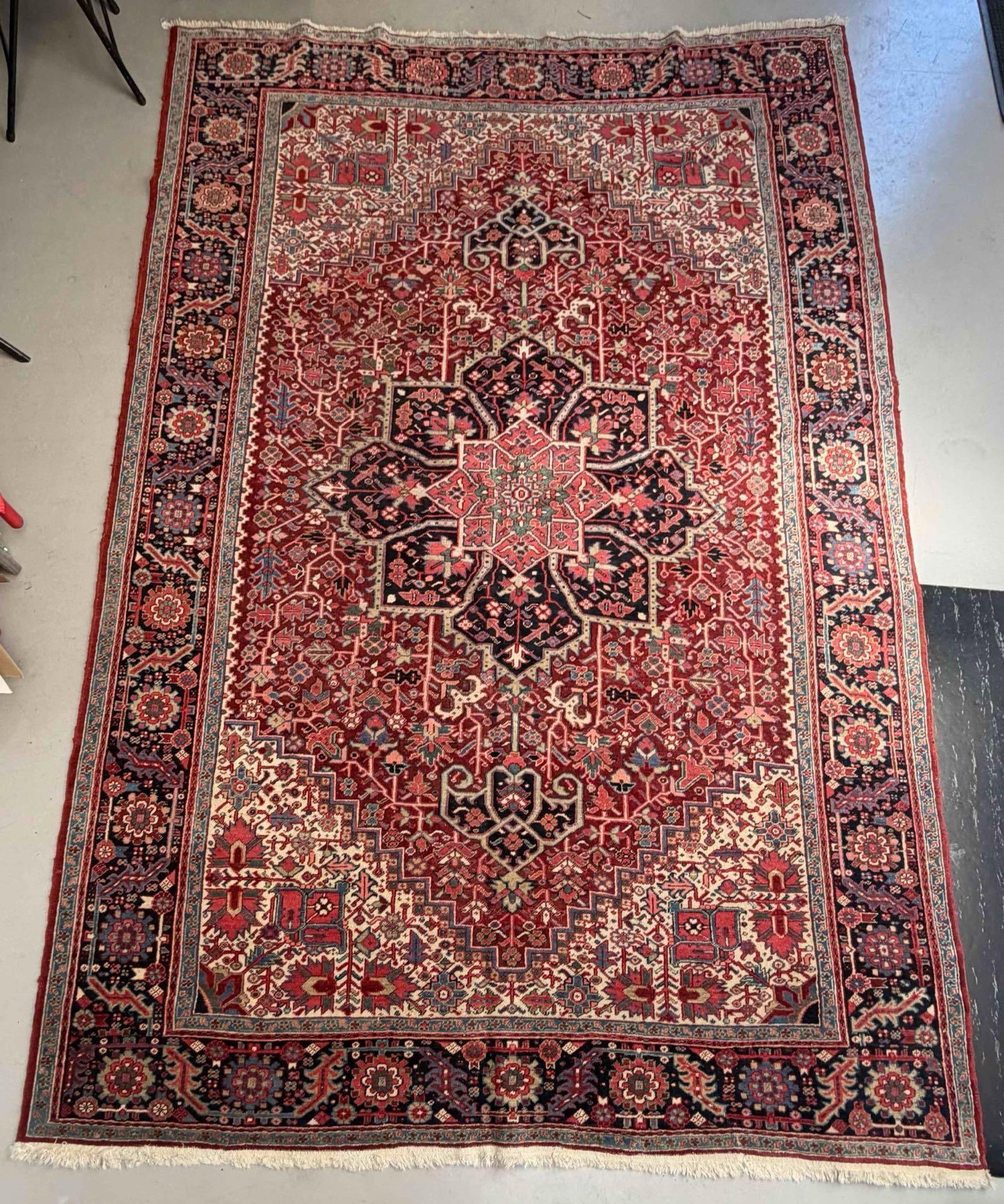 Persian Heriz Rug: Persian Heriz Rug. Hand-knotted wool carpet with a red field centered by a large dark blue geometric medallion, surrounded by angular vines, stylized floral motifs, and ivory corner spandrels. Approxi