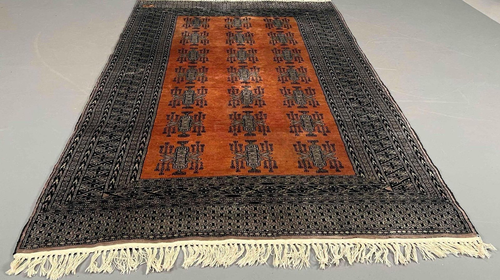 Bokhara Style Area Rug With Inscription: Bokhara style wool area rug, with repeating medallions on a rust-orange field, enclosed by multiple geometric borders in black and ivory tones. One border with woven Arabic-script signature or inscrip
