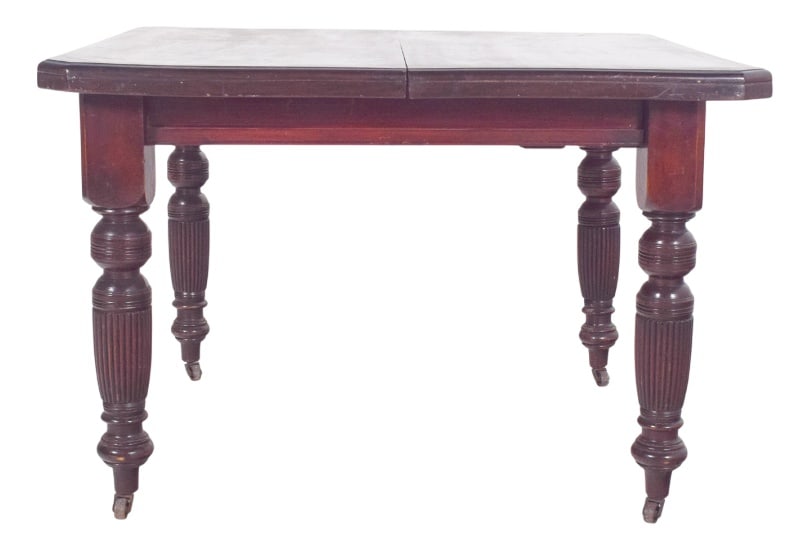 English Victorian Dining Table, 1890s: English dining table. It has a square top with slightly jagged edges, a smooth surface and a darkened finish. It rests on four turned baluster legs decorated with vertical fluting with small metal whe