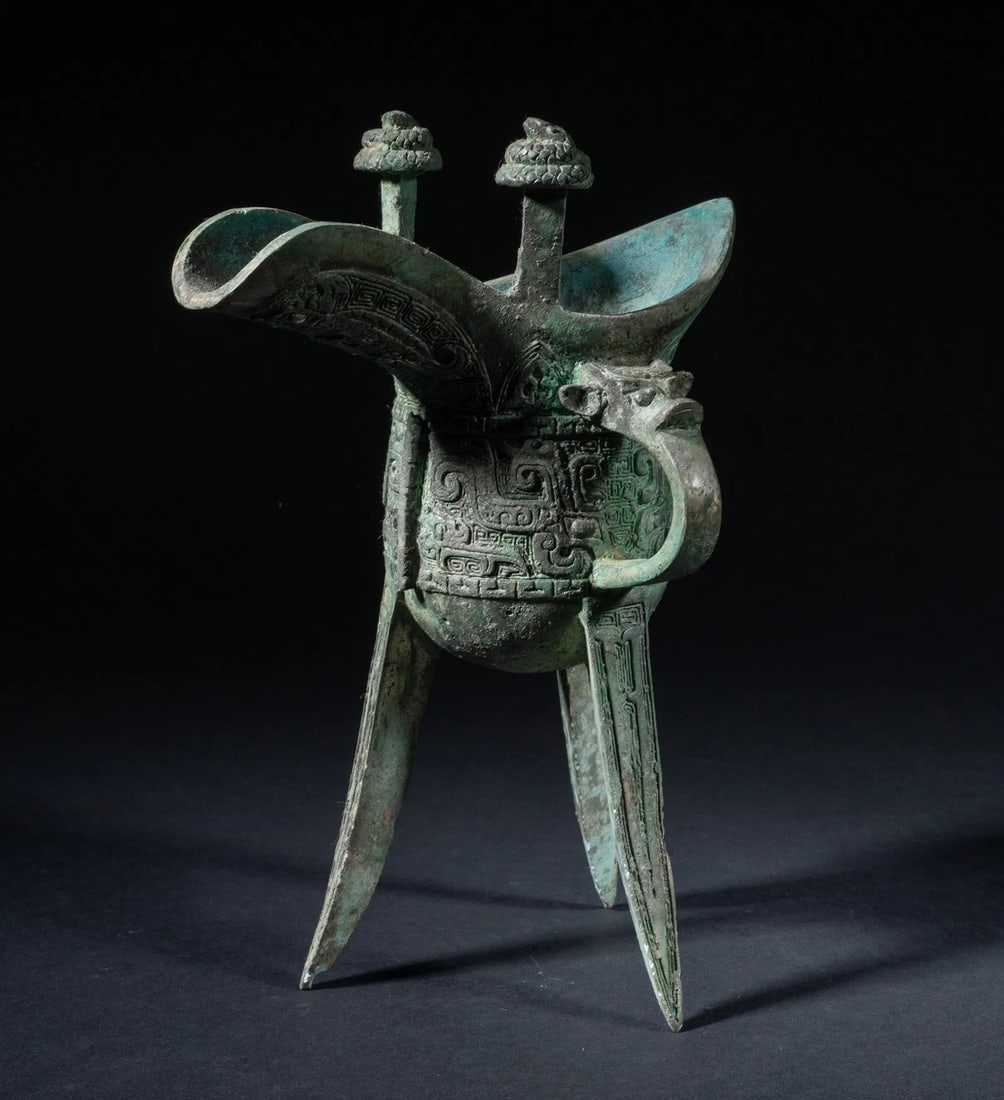 Chinese Antique Archaic Bronze Tripod Wine Vessel (Jue) with Taotie Mask Decoration, Shang Dynasty: Supported on three splayed, blade-like legs, the body with a mythical beast-head handle on one side, elongating at the rim to a cupped spout at one end and a raised pointed tip at the other, the rim t