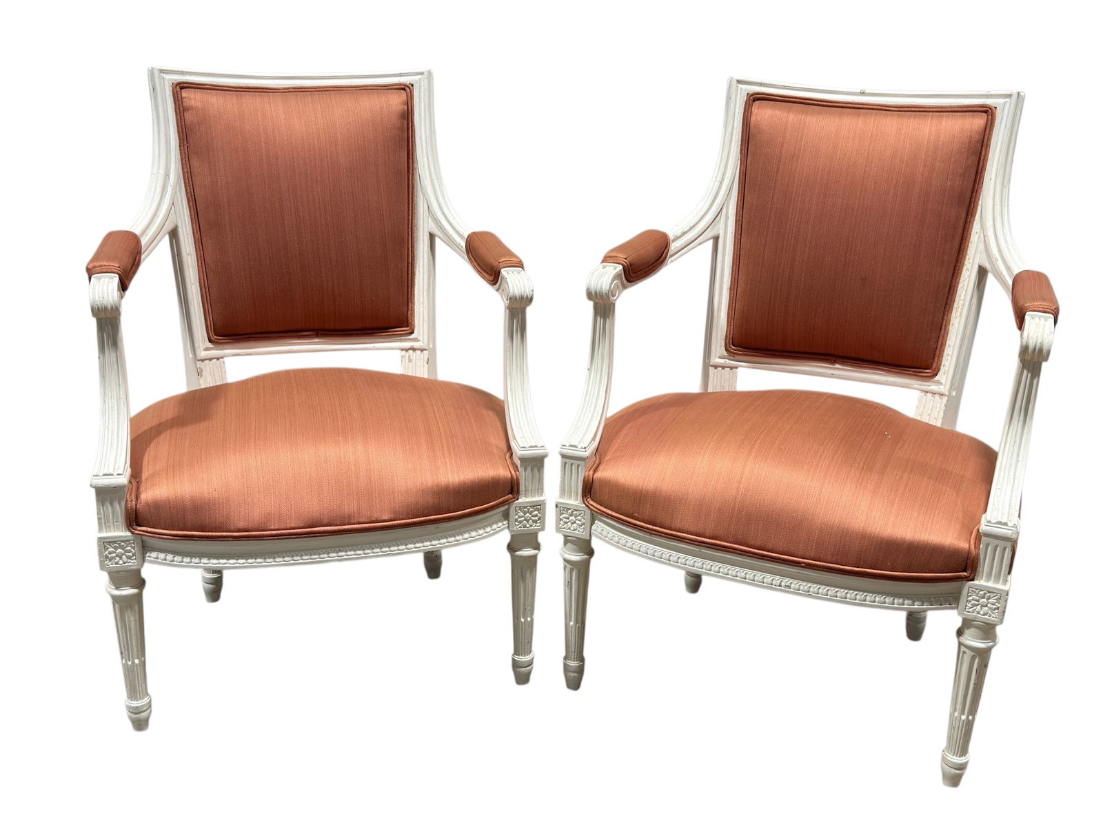 Pair of French Painted Louis XVI Style Armchairs: Elegant pair of French Louis XVI style armchairs featuring carved and painted frames with fluted legs, neoclassical rosette detailing, and gently curved arms. Upholstered in a warm peach-toned fabric