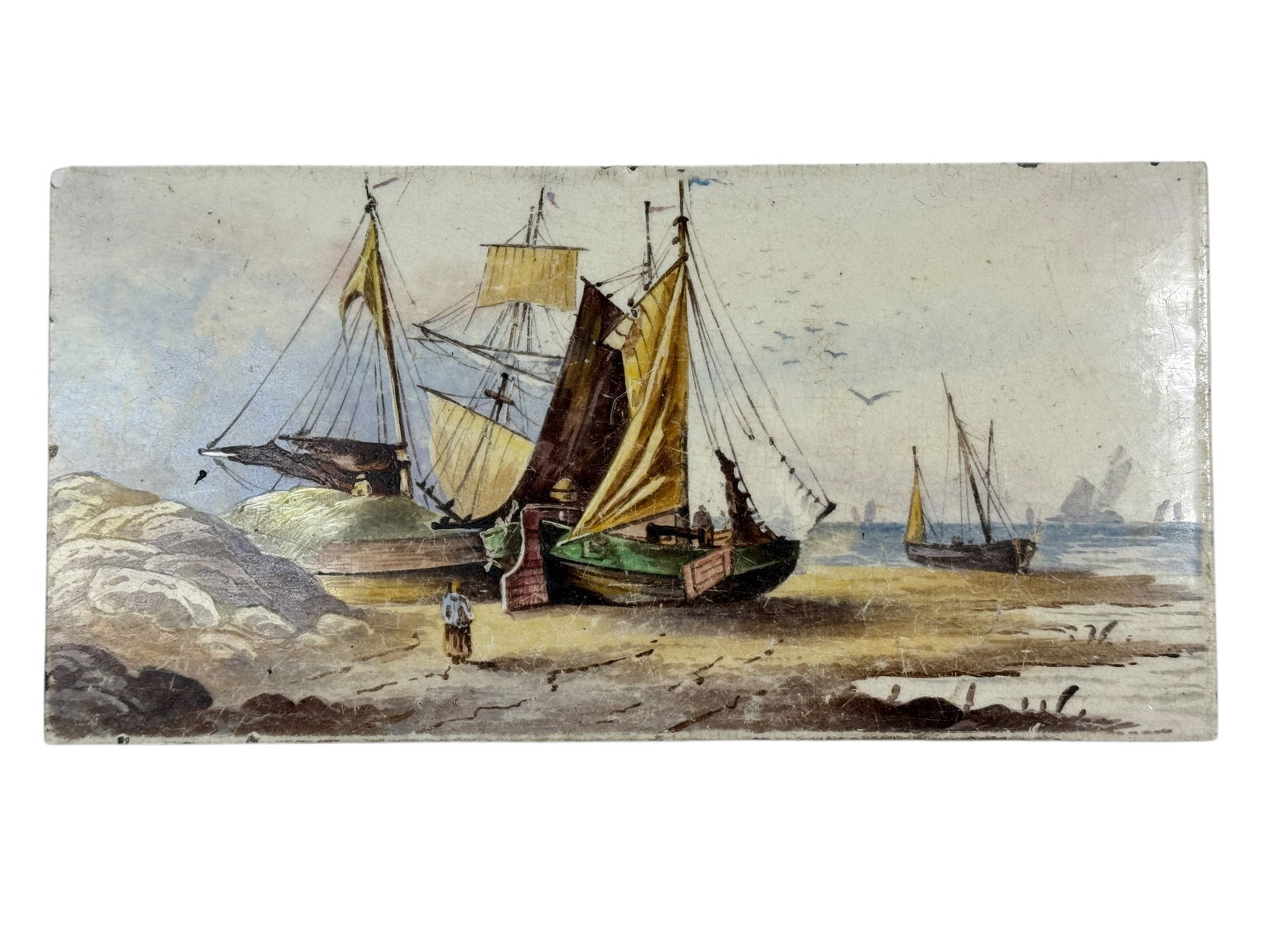 19th C. Hand-Painted Maritime Tile w/ Fishing Vessels: This hand-painted ceramic tile portrays a coastal shoreline scene with fishing boats resting on land rather than underway at sea. The vessels are shown beached along the shore with sails raised or fur
