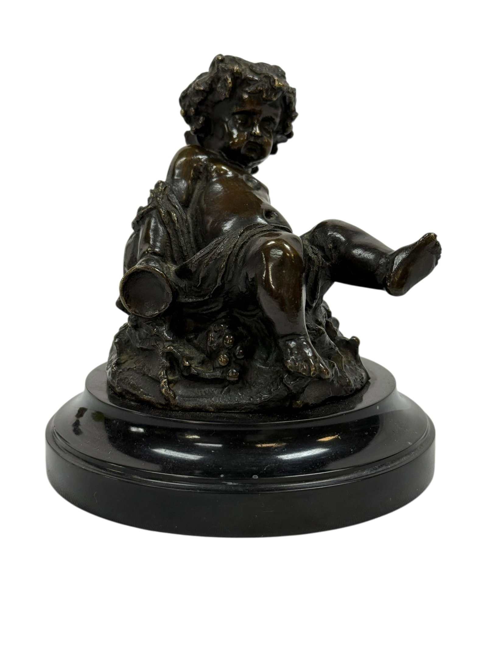 After Clodion Bronze Putti Figure on Marble Base: Charming bronze sculpture in the manner of Clodion (Claude Michel), depicting a playful putto seated atop a naturalistic base with draped cloth and a small vessel at his side, the figure rendered with