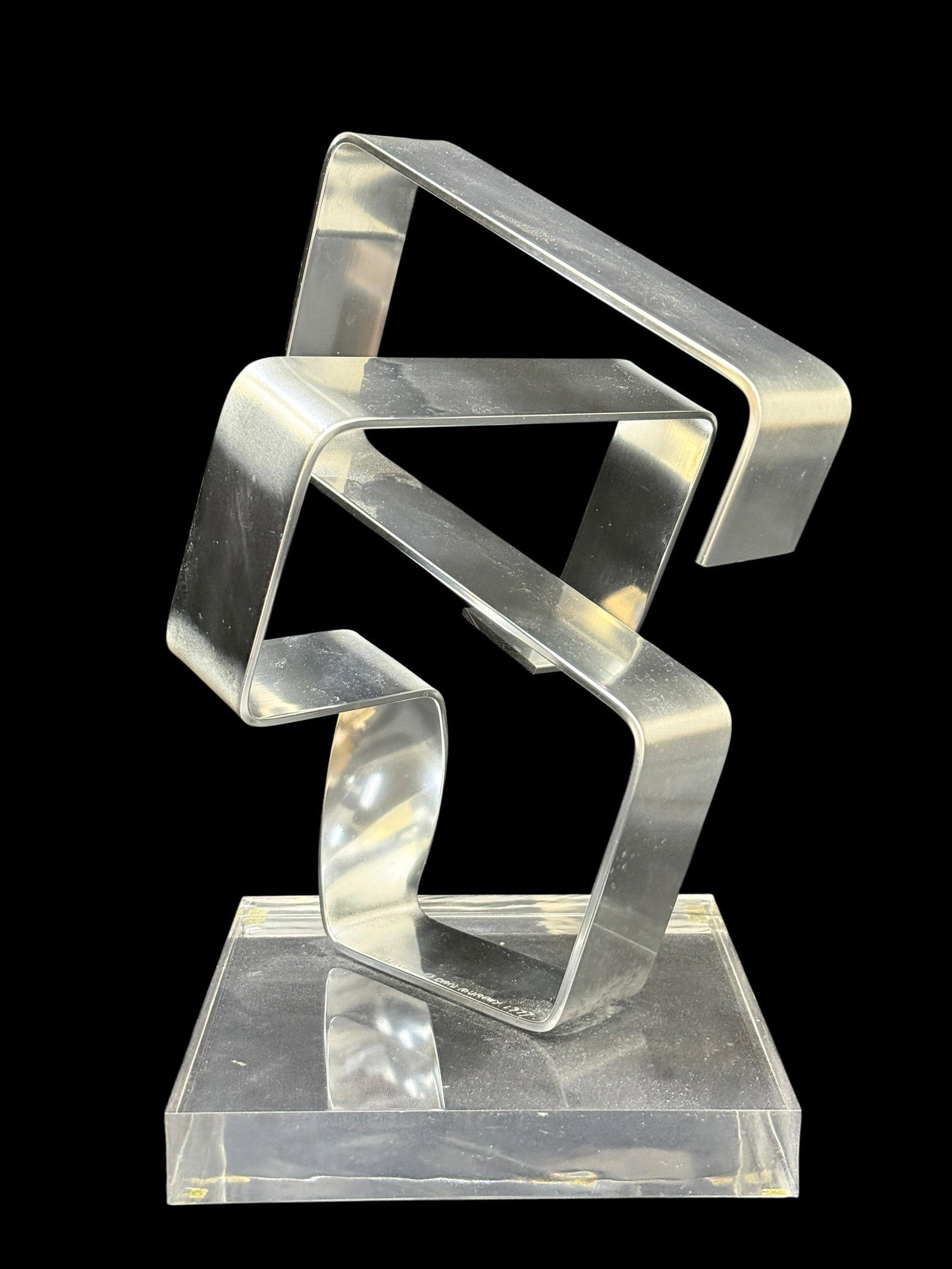 1977 Dan Murphy Abstract Metal Sculpture on Lucite Base: A striking 1977 modernist abstract sculpture titled “Justice,” executed in brushed aluminum with interlocking geometric ribbon-like forms surrounding a central cylindrical element, mounted on 