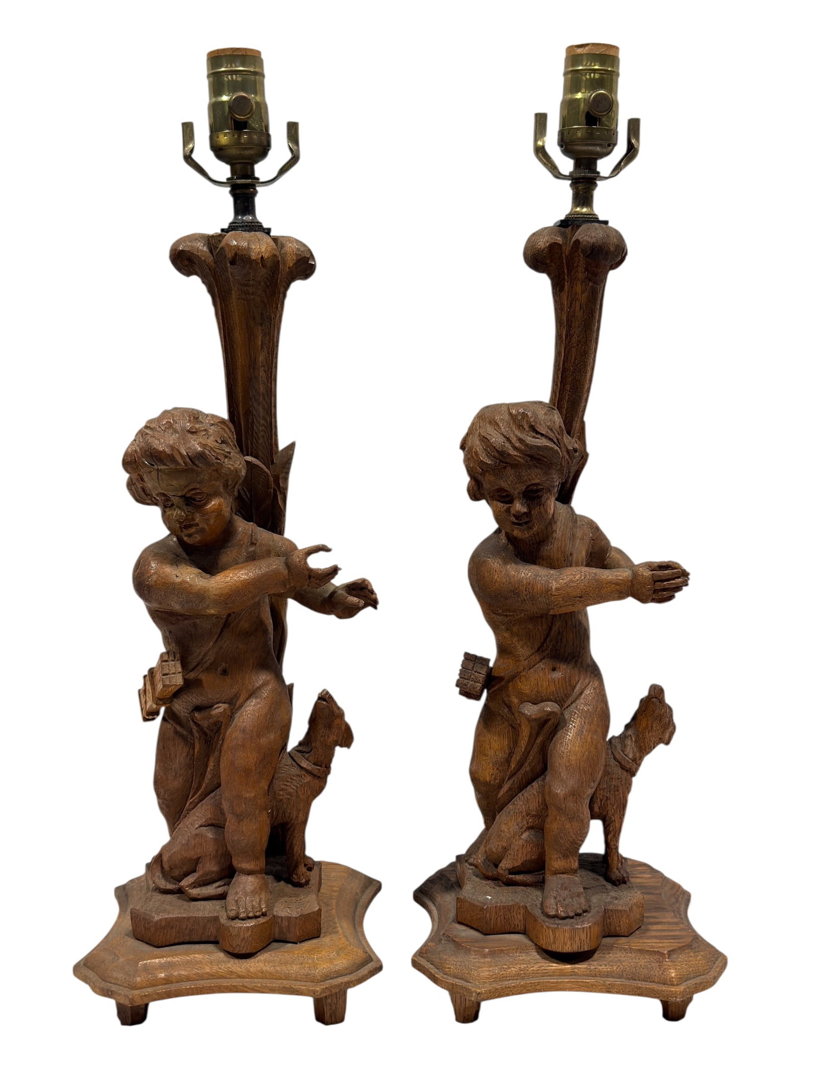 Pair of Carved Wood Putti & Dog Figural Table Lamps: This pair of lamps features a sculptural wooden base depicting a young child, styled similarly to traditional putti or cherubs, standing beside a small canine companion. The figures are positioned aga