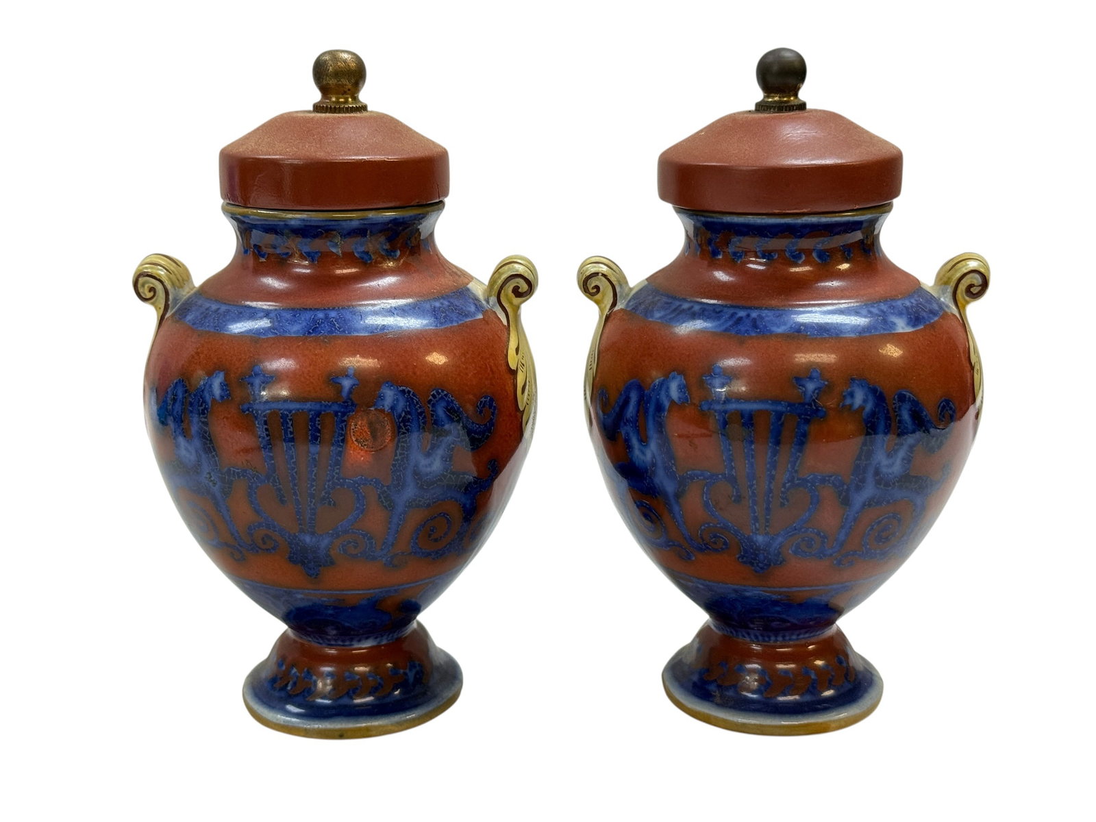 Pair of Small Neoclassical Style Lidded Ceramic Urns: A decorative pair of Neoclassical-style ceramic urns featuring a rich oxblood-red ground accented with cobalt blue decoration, including classical lyres, scrolling motifs, and stylized horses, with