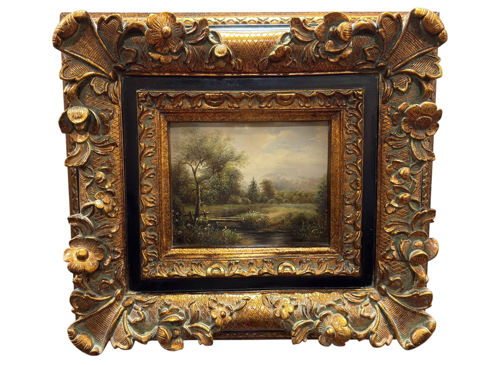 Early 20th Century Pastoral Landscape Oil on Board in Elaborate Floral Gilt Frame: Oil on board pastoral landscape signed at the lower right, depicting a tranquil wooded meadow with a narrow reflective stream, rustic footbridge, flowering grasses, and dense vegetation receding into 