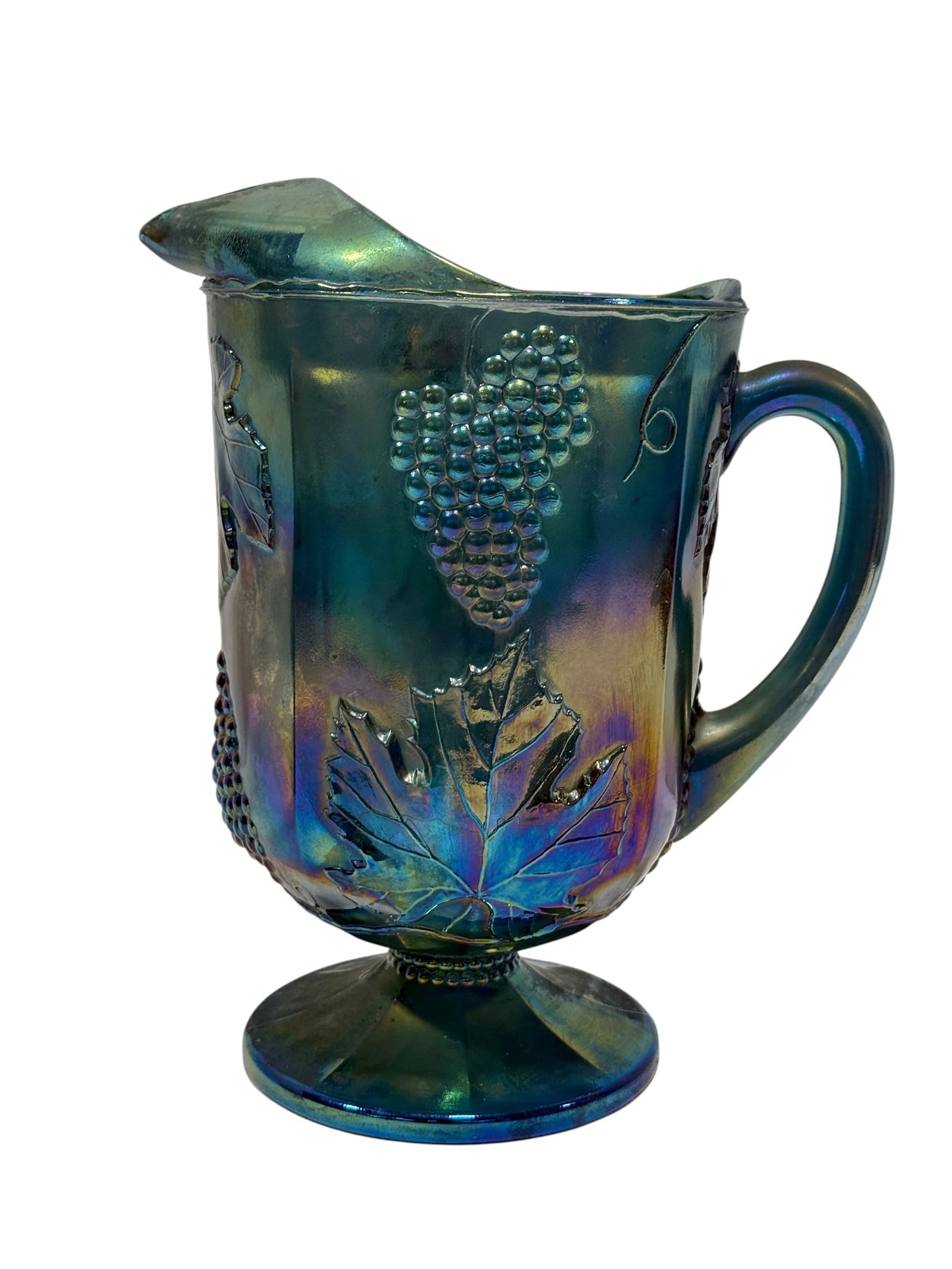 Early 20th C. Blue Iridescent Carnival Glass Grape Pattern Pitcher: A striking early 20th century carnival glass pitcher formed in deep blue-green glass and richly iridized with shifting teal, cobalt, violet, gold, and emerald highlights. The tall paneled body is deco