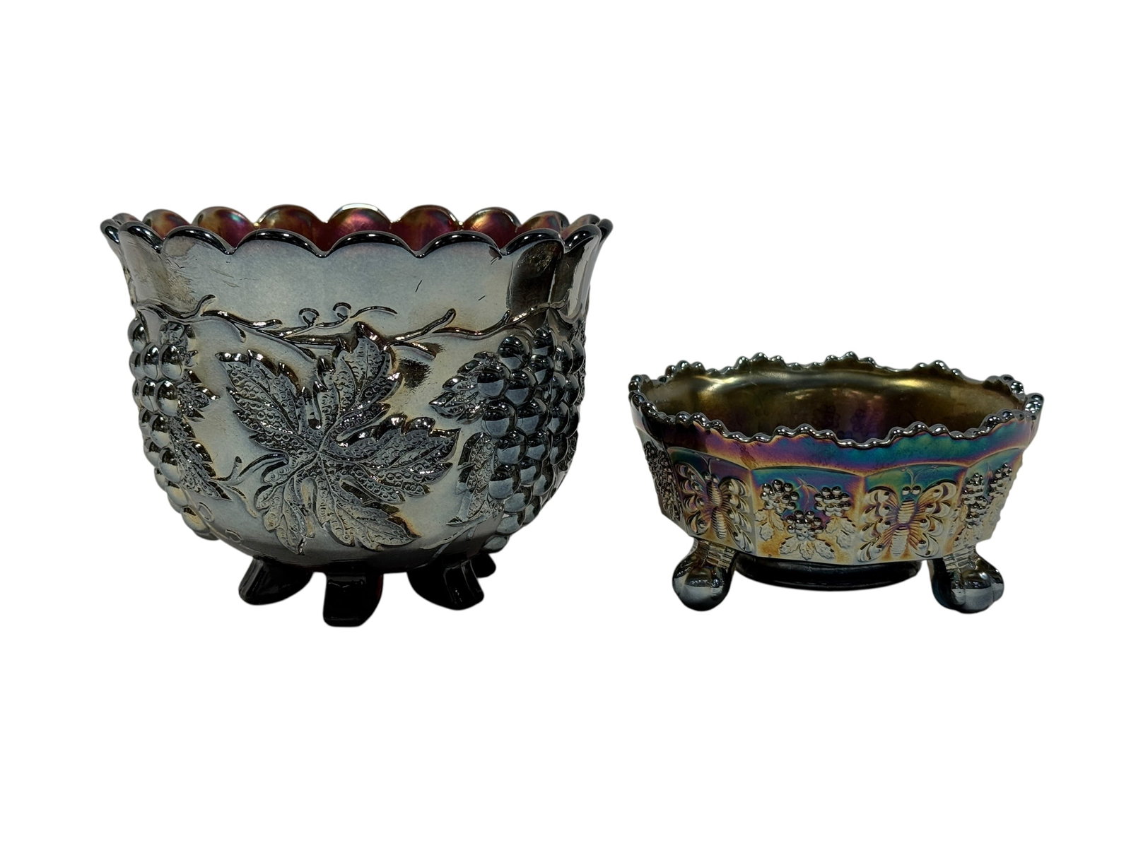 Pair of Vintage Iridescent Carnival Glass Footed Bowls: A decorative pair of vintage iridescent carnival glass bowls with rich silver, gold, violet, blue, and green surface color. The larger bowl is molded with bold grape clusters, broad leaves, and curlin
