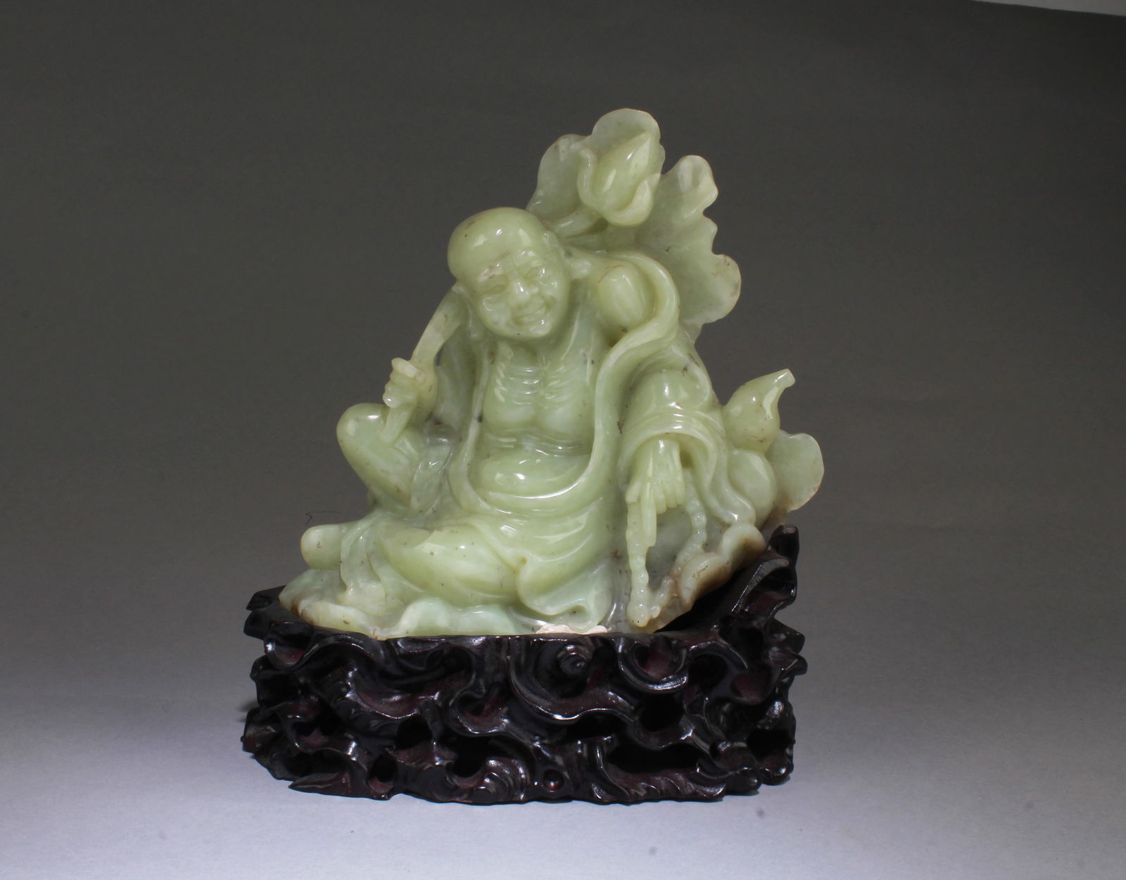 Chinese Jade Carved Ornament: Intricately carved jade ornament, depicting a smiling buddha wearing flowing robe in sitting position, holding a large lotus leaf on one hand and prayer beads on the other hand. Comes fitted with a