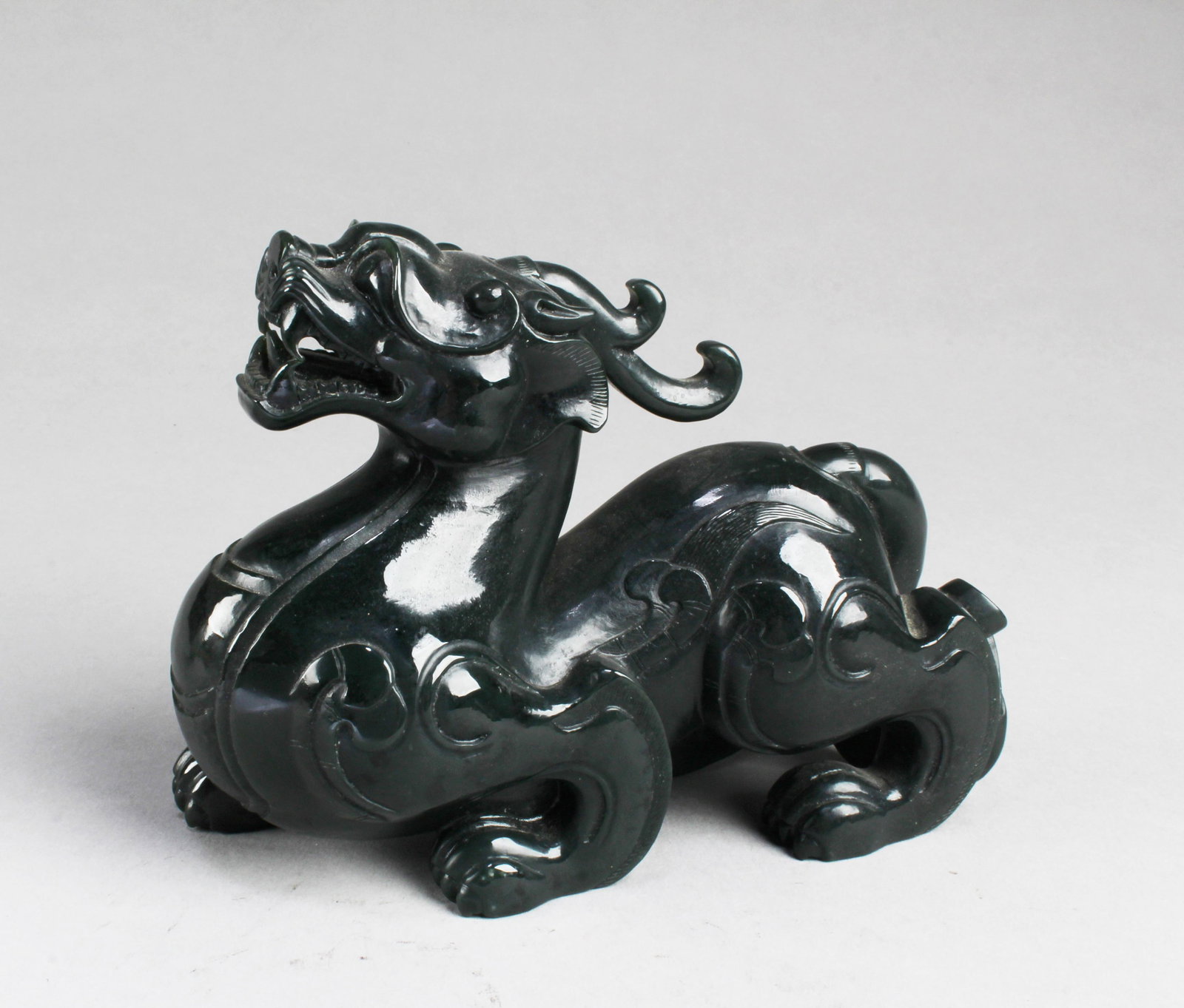 Chinese Spinach Green Nephrite Jade Ornament: Chinese Spinach Green Jade Carved Ornament, depicting a intricately carved mythical creature, showing it's mouth, teeth, mane, in sitting position. Length: 6.25 "
