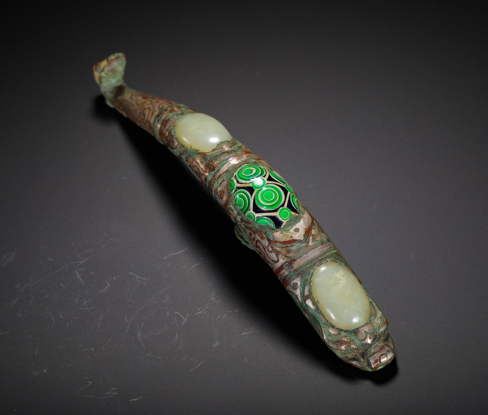 SILVER INLAID WITH GLAZED WHITE JADE AND COPPER HOOK FROM THE WARRING STATES PERIOD: L:19.5cm (7.68in) Weight:220g (0.49lb)A Warring States bronze belt hook inlaid with silver, white jade and glass eye bead. Designed with beast head terminal, the bronze body is engraved with silver in