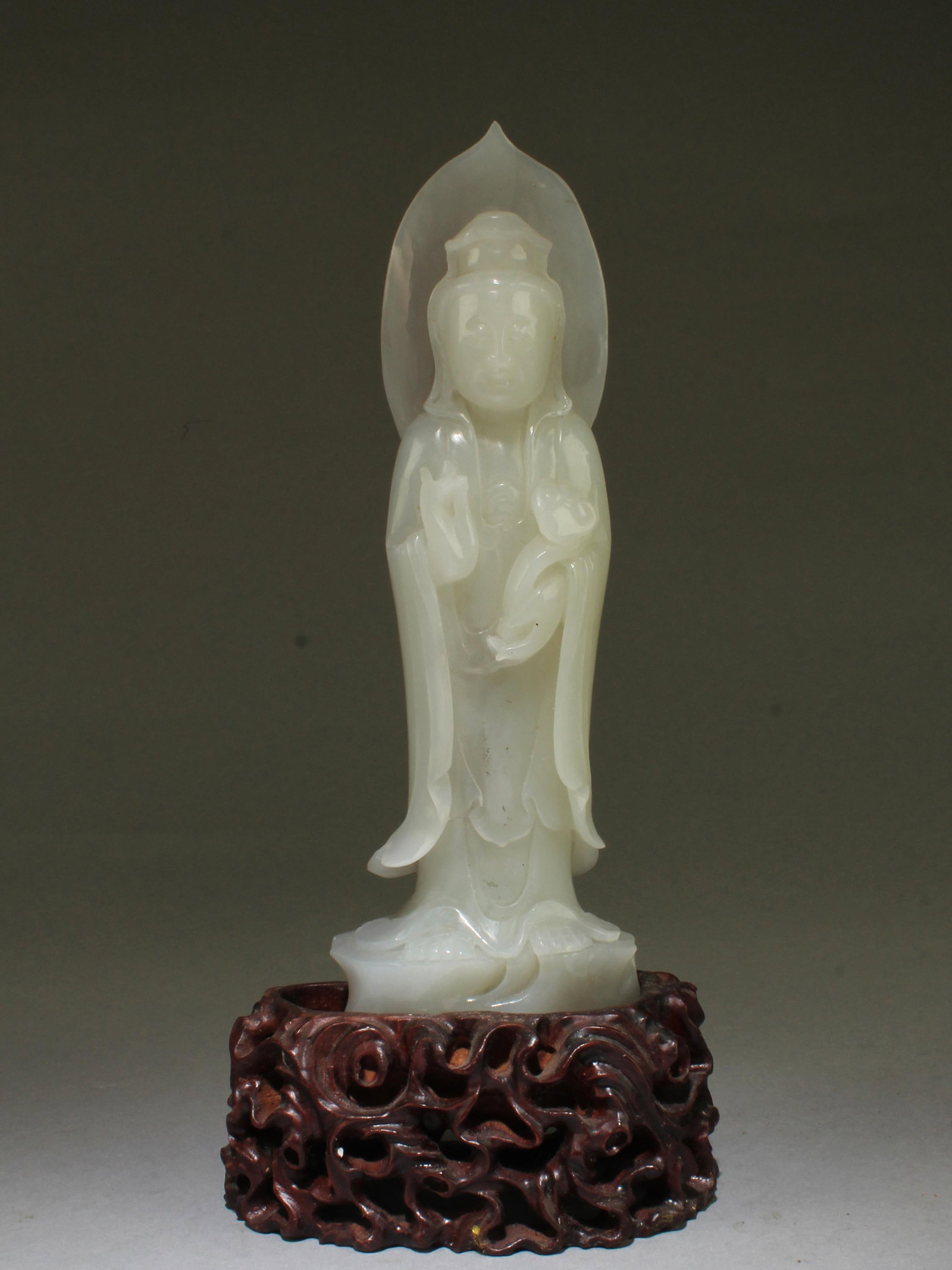 Chinese Jade Carved Guanyin Statue: Intricately Carved Jade statue, depicting a guanyin standing on a throne, left hand holding a ruyi sceoter, right hand in mudra, wearing flowing robe. Comes fitted with a wooden stand. Height: 7.25"