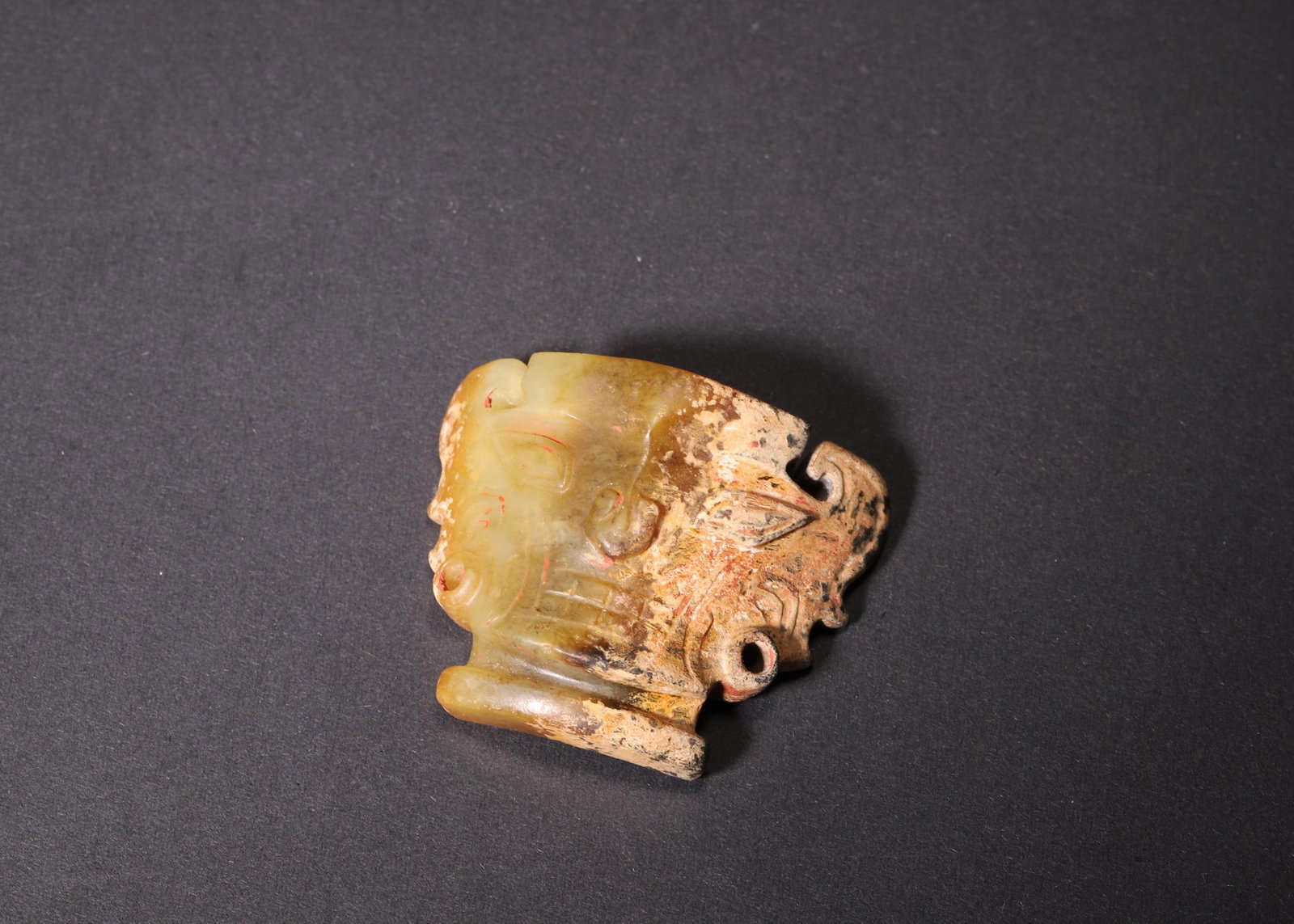 A Jade Human-Mask Pendant, Shang Dynasty: Wt 38.0g (0.08 lb); L 4.5cm (1.77 in); W 3.5cm (1.38 in); H 2.0cm (0.79 in)A Shang Dynasty jade human-mask pendant. Carved with a solemn and stylized human face, it features natural patina and cinnaba