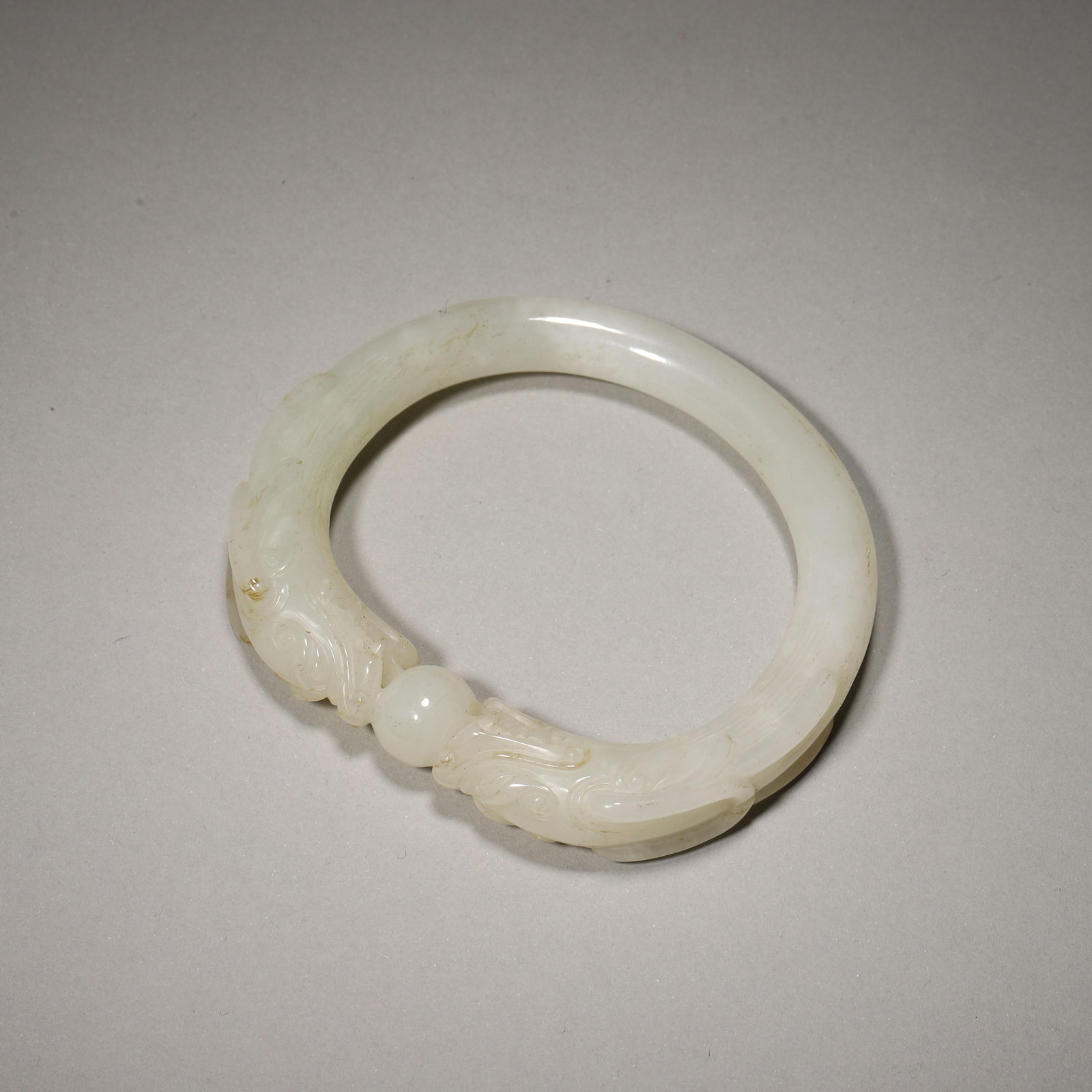 A HETIAN WHITE JADE BANGLE CARVED WITH DOUBLE DRAGONS CHASING PEARL, QING DYNASTY: Inner D 59.00mm (2.32 in); Wt 59.00g (0.13 lb),Carved from single piece of Hetian white jade with soft greasy luster and faint russet natural inclusions. Two confronting dragon heads at one end clasp 