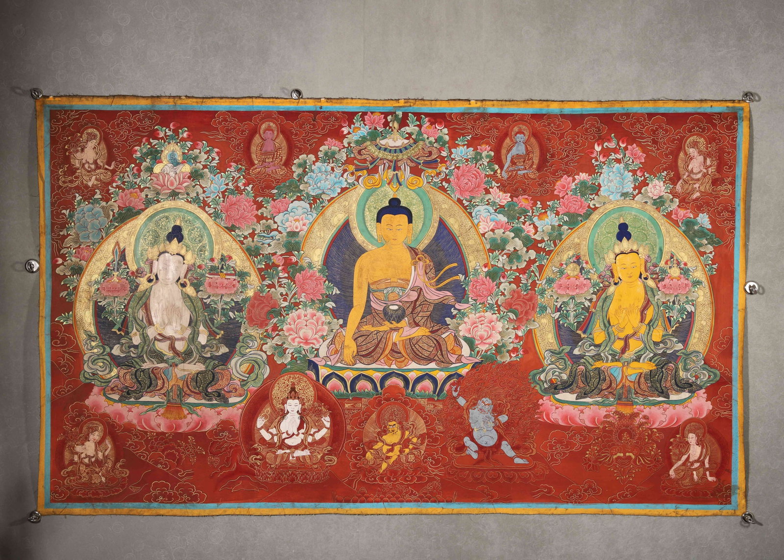 TIBETAN BUDDHIST THANGKA OF THE THREE BUDDHAS, 19TH CENTURY: 60.24in x 35.43in (153.0cm x 90.0cm)This 19th-century Tibetan Buddhist Thangka depicts the Three Buddhas with vivid mineral pigments and gilded details. The rich red background, solemn deities, and in