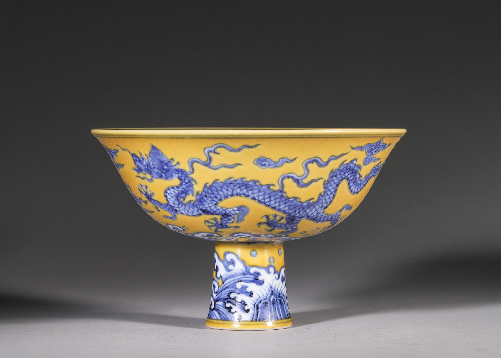A YELLOW-GROUND BLUE AND WHITE STEM BOWL WITH DRAGON AND WAVES, XUANDE PERIOD MING DYNASTY: L 17cm (6.69 in); H 10.5cm (4.13 in),A Ming Xuande stem bowl with wide flared rim, covered in soft yellow ground glaze. The exterior is painted in rich cobalt blue with two vigorous dragons chasing fl