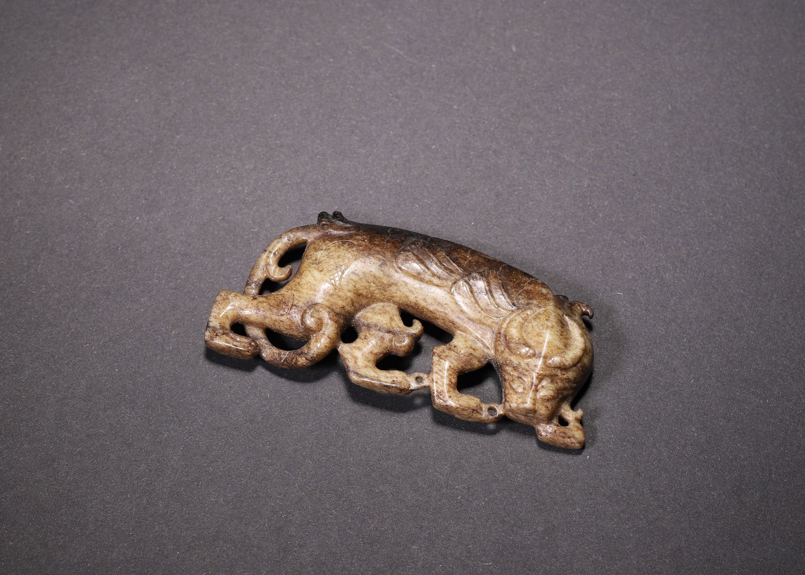 A Jade Auspicious Beast Pendant, Han Dynasty: L 7.0cm (2.76 in); W 3.0cm (1.18 in); Wt 29.0g (0.06 lb)A Han Dynasty jade pendant carved from Hetian jade, showing deep brown and beige patina. It depicts an auspicious beast in a crawling posture wi