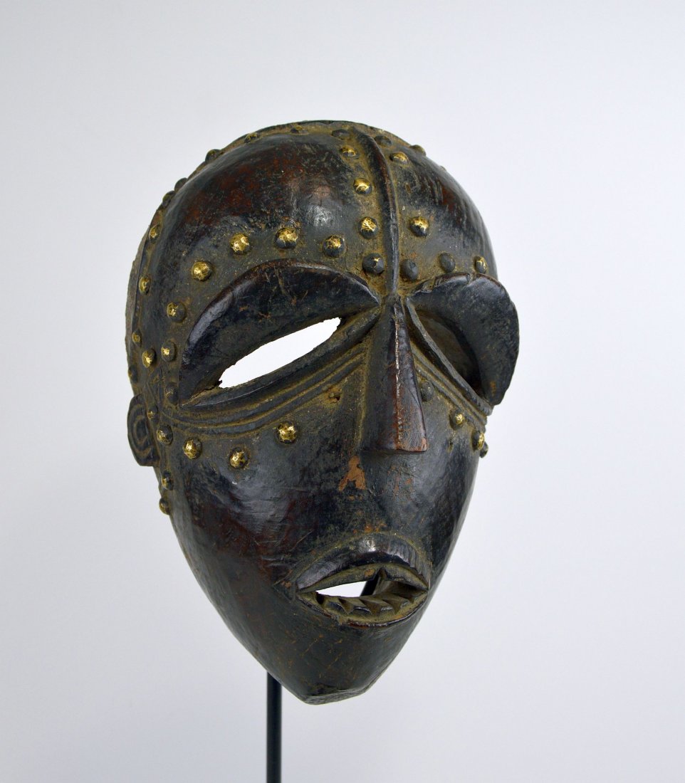 A Fine Old Bete African Mask: Carved wooden mask with stylized facial treatment and a well handled wooden surface patina, accented by metal tacks. The mask shows heavy wear and use.The mask itself measures 13 inches tall. The mask
