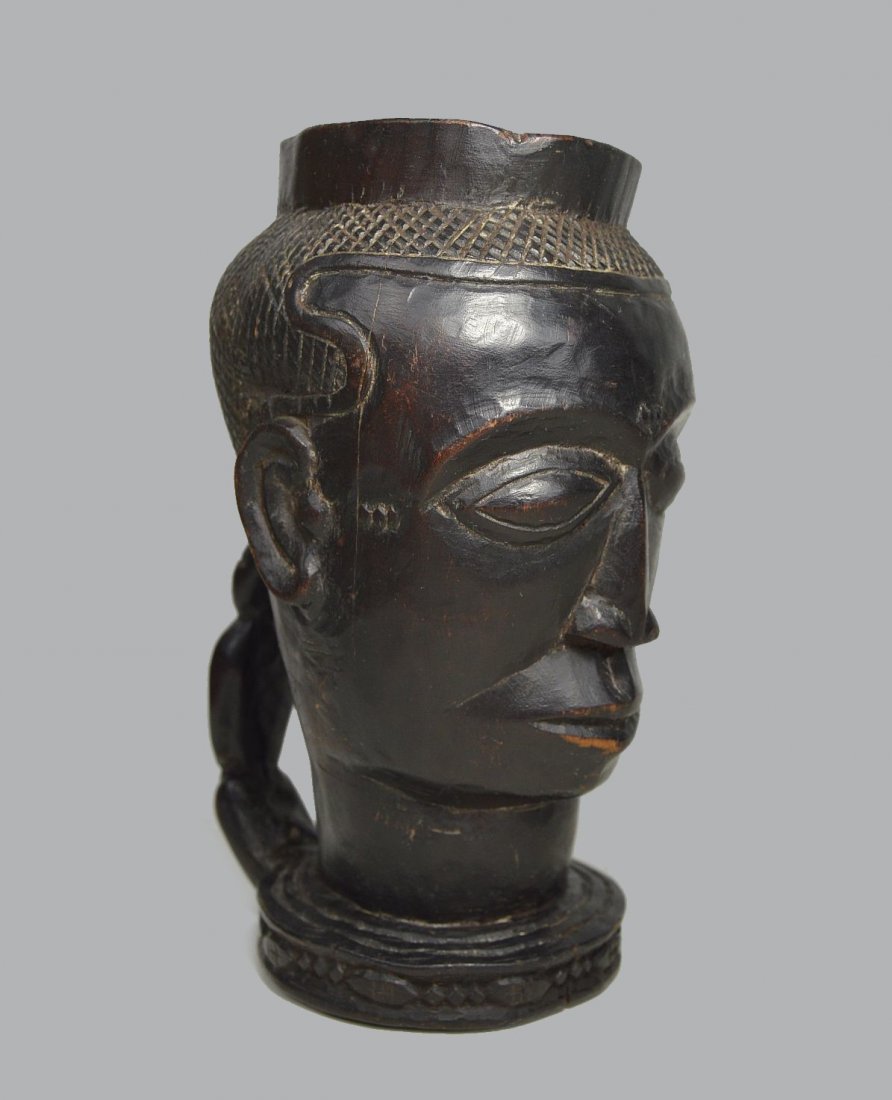 KUBA PALM WINE CUP WITH ANCESTOR FACE: A finely crafted wooden palm wine beaker with handsome ancestor face and carved handle, all from a single piece of wood. Kuba peoples, Congo, Africa. The kuba cup measures 8 inches tall.