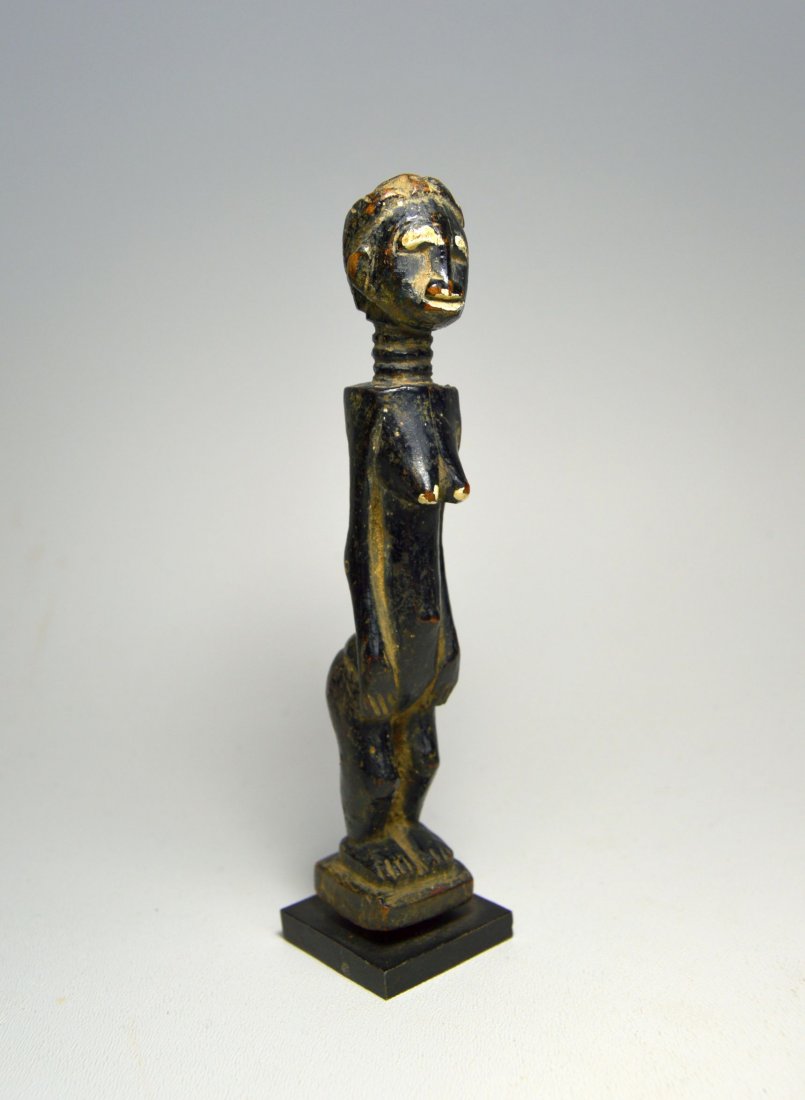 LOVELY OLD BAULE PAINTED MINIATURE FEMALE IDOL: A lovely old carved wood and painted female effigy with stylized coiffure,ringed neck, hands held down along the torso, protruding navel and accented breast. Obvious weathering and handling suggest ri