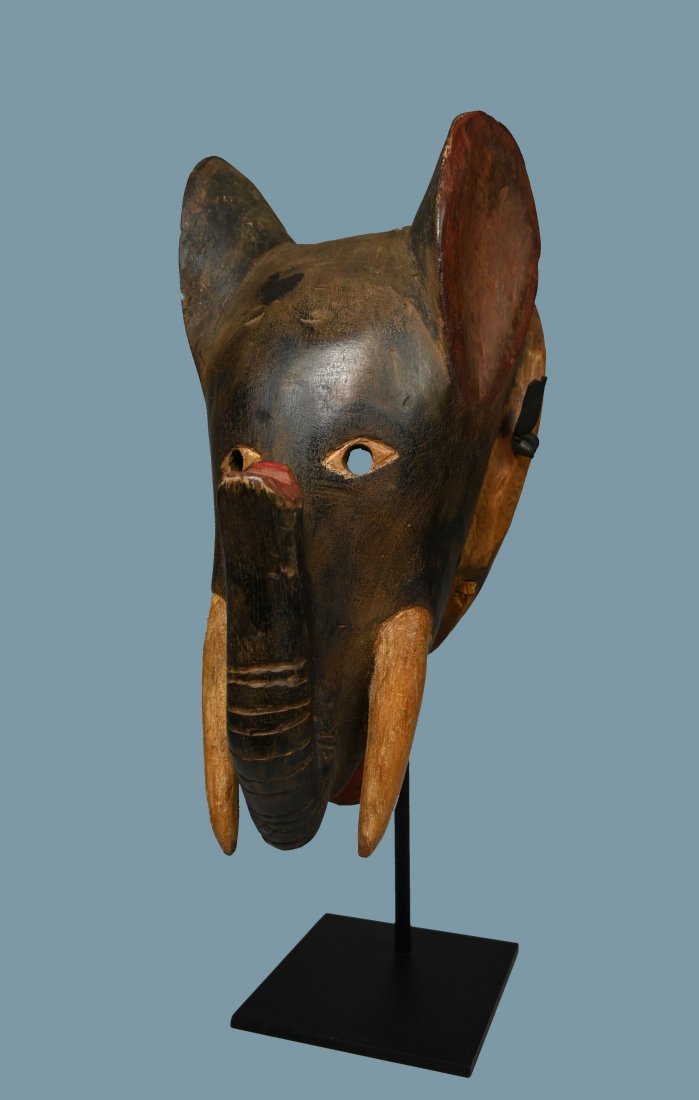 A Baule Elephant African mask: A carved wooden mask in the form of an Elephant, with tusks and large ears, painted black. Baule peoples, Ivory Coast, West Africa. The mask measures 13 x 6 inches. A metal display stand can be includ