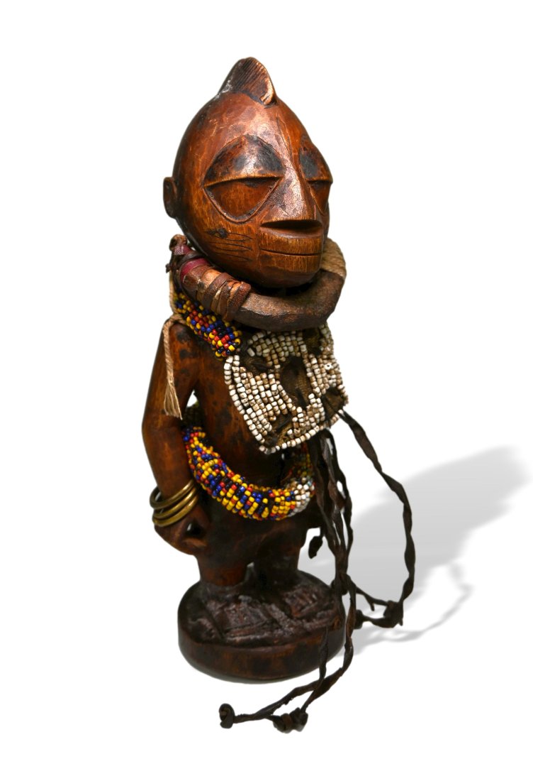 A Yoruba Female Sculpture: Carved wooden effigy embellished with numerous collars, bands and skirt of beads and textiles, along with hammered bronze bracelets. The figure has classical facial features and some unusual organic s