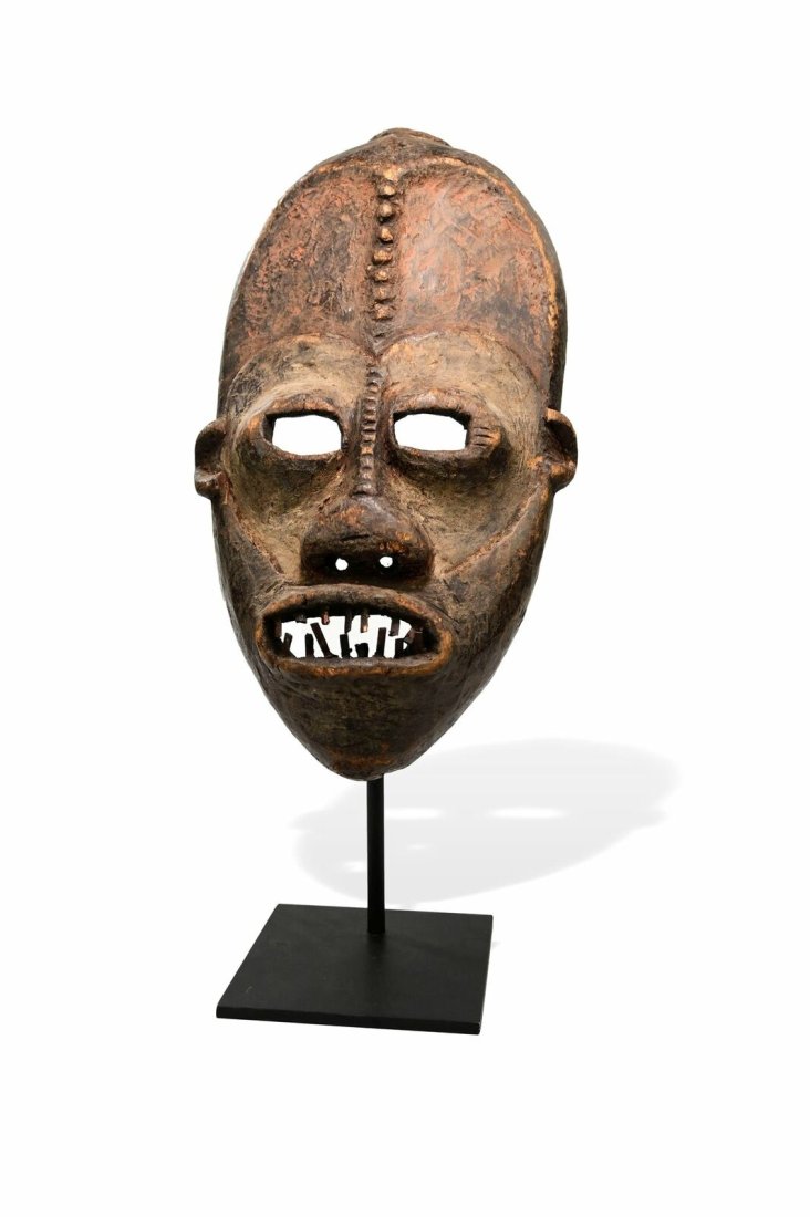 A Ngbaka African Mask: A dramatic carved wooden mask with exaggerated facial features, raised scar designs on the brow of the nose as well as inset crooked teeth. The mask face has been accented with kaolin clay pigments. A