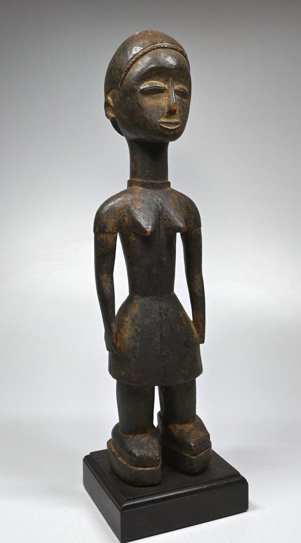 A Serene Baule Akan Sculpture: A charming carved wooden female effigy with a serene face and well handled and worn surface. The figure is shown in western dress and wearing sandals. Baule peoples, Ivory Coast, West Africa. The figu