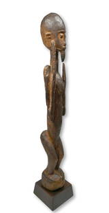 A Dogon Nommo Sculpture Ex Joan Didion Estate: A Tall slender carved wooden shrine idol, likely a 19th century example or earlier. The sculpture is shown with hands to the air in an act of prayer to the star sirius, possibly for rain. There is a N