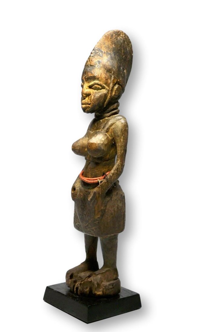 A Yoruba Female Sculpture Ex Banks Collection: A carved wooden female figure from the Yoruba peoples, perhaps a colonial era example shown wearing western bra/top and skirt, with a well handled , weathered surface. This sculpture measures 14.5 inc