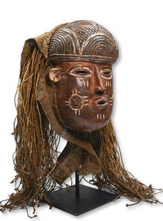 A Chokwe Pwo African mask: A carved wooden dance mask with incised facial scars and tribal motifs, accented with kaolin clay pigments, and adorned with an elaborate woven grass hair coiffure and netting for the dancer to wear.