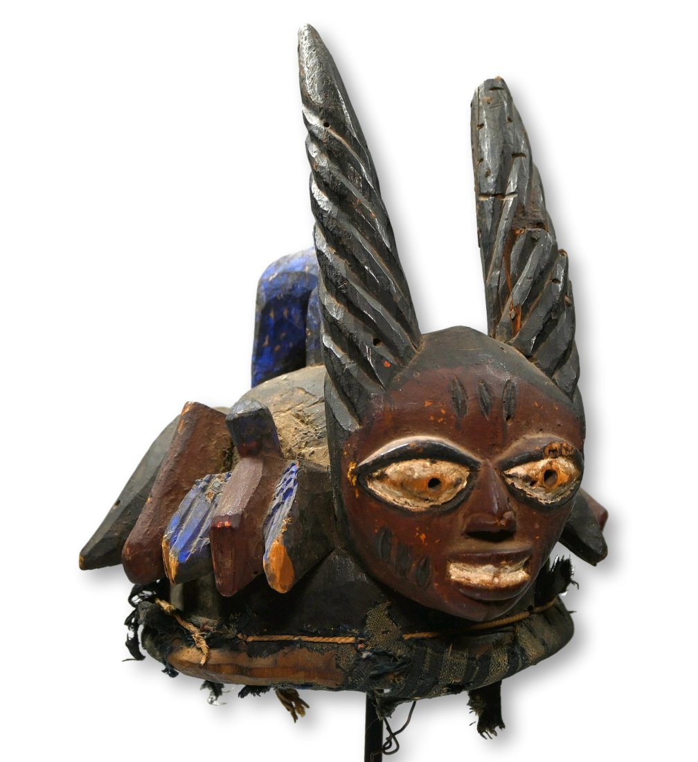 A Yoruba Egungun Mask with Avian Images: Carved wooden cap mask with stylized anthropomorphic head on top with tall twisted horns and painted with organic pigments. The cap mask features several zoomorphic and mask like faces , the head of t